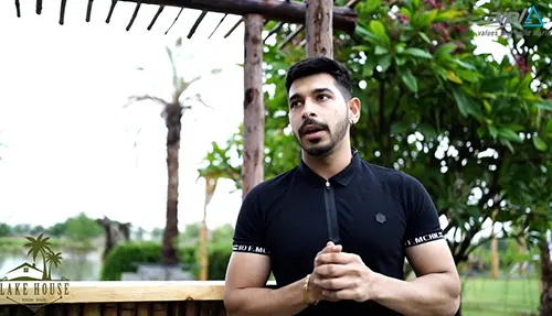 Man stands outdoors at AIPL DreamCity township in Ludhiana, Punjab. Tropical plants and pergola visible in background.